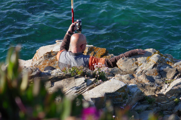 Bald man fishing with fishing pole rod from cliffs rocks. Flowers stock photo
