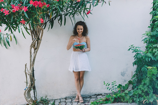 Tourist woman on vacation standing under flowering tree and eating sliced watermelon with bamboo spoon
