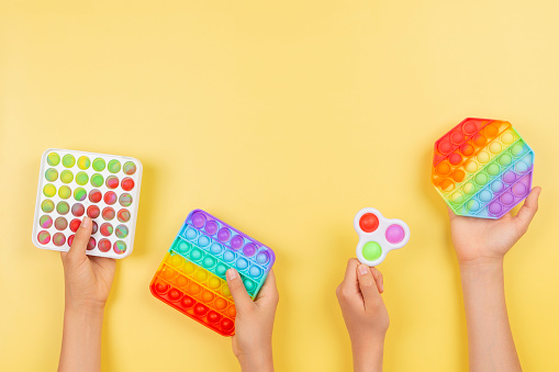 Many hands holding pop it fidget toys on yellow background. Push pop-it fidgeting game helps relieve stress, anxiety, autism, provide sensory and tactile experience for children