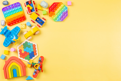 Baby kids toys background with wooden rainbow house, train, car, plane, pop it fidget toys and colorful blocks on yellow background. Top view, flat lay
