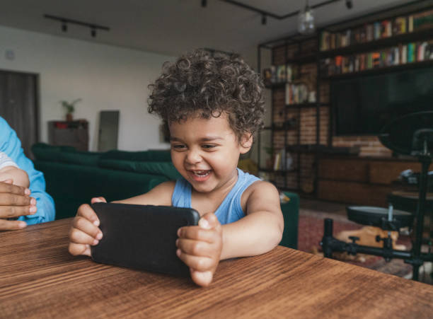 menino negro de 3 anos assistindo desenhos animados em smartphone sentado à mesa de jantar - 2 3 years children only group of people enjoyment - fotografias e filmes do acervo