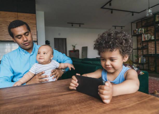 menino negro de 3 anos assistindo desenhos animados em smartphone sentado à mesa de jantar - 2 3 years children only group of people enjoyment - fotografias e filmes do acervo