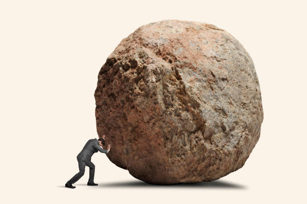 Man Attempts To Push Giant Boulde A man leans into his effort to push a giant round boulder. boulder rock stock pictures, royalty-free photos & images