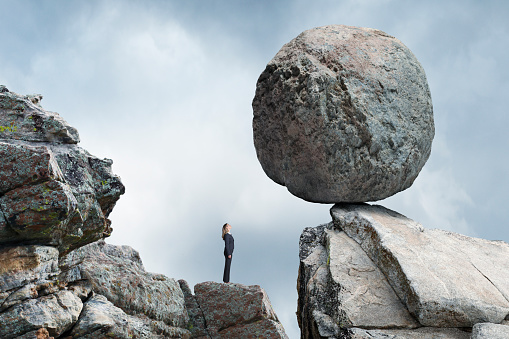 A businesswoman stands and looks up a a large round boulder that is precariously balanced on. the rocky promontory above her.