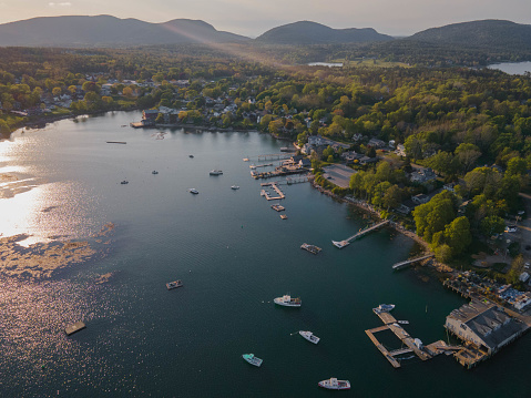 A variety of boats, from small fishing boats to large sailboats, line the docks in the harbor. The sun rays shined across the water just before starting to set behind the mountains in the distance.