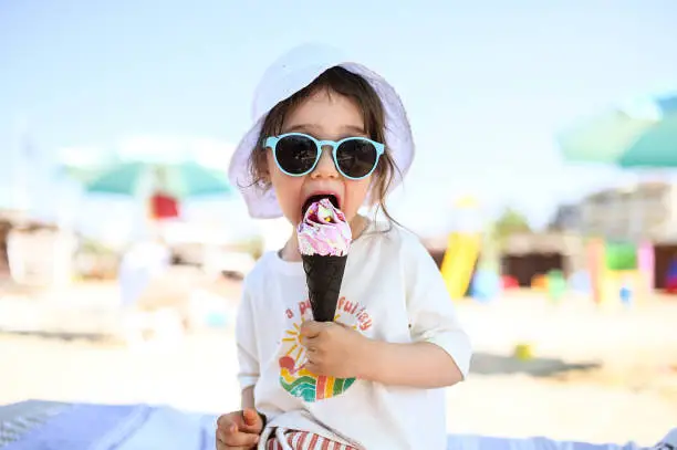 Funny caucasian little girl 2-3 years old eating cone with ice cream on the beach. Summertime concept. Family vacation and holidays concept. Selective focus. Child with sunglasses.