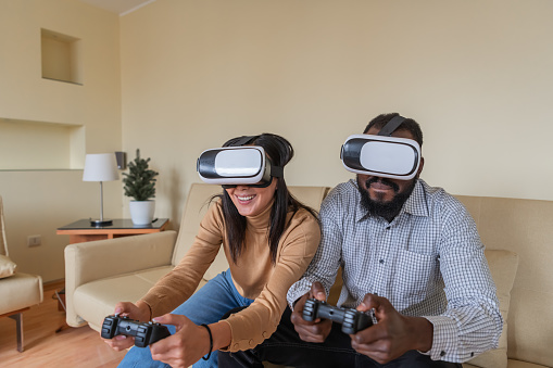 Smiling Young Man is Playing Computer Games with his Excited Beautiful Girlfriend of Chinese Ethnicity. Smiling Young Multi-Ethnic Couple is Enjoying 3D Games in Virtual Reality with Goggles and Joysticks.