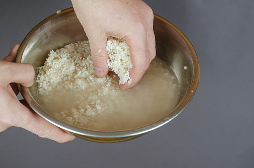 A man washes raw white rice in fresh water. Hands stirring grains of uncooked wet rice in a metal bowl. Food, cooking. Close-up, selective focus.