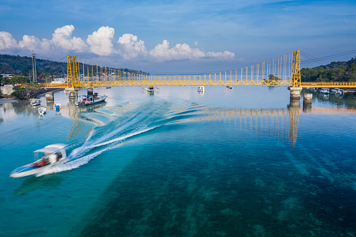 Aerial drone view of a local boat passing under the yellow bridge that is connecting the island of Nusa Lembongan and Ceningan in Bali Indonesia.