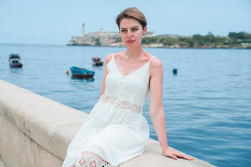 Young woman wearing a white dress, sitting by the ocean