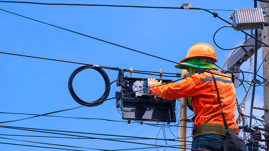 Rear view of technician on wooden ladder is working to install fiber optic and splitter box on electric pole against blue sky background