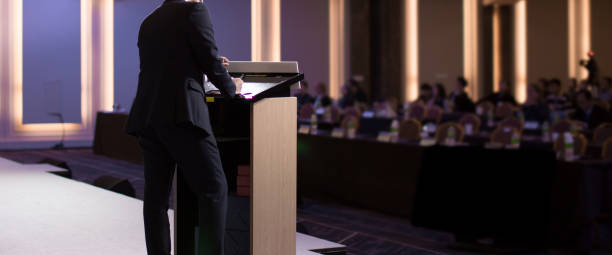 istock Conference photo audience and speaker giving speech. Seminar presenter on a panel during forum. Corporate manager in sales executive training discussion on stage. Investor pitch presentation. 1324999257