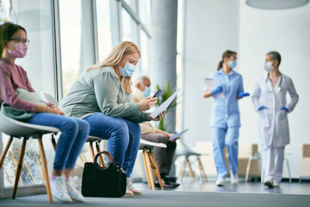 Woman with face mask using smart phone while reading her medical report in waiting room at the clinic. Female patient examining medical paperwork and using mobile phone while sitting in hallway at the hospital. waiting room stock pictures, royalty-free photos & images