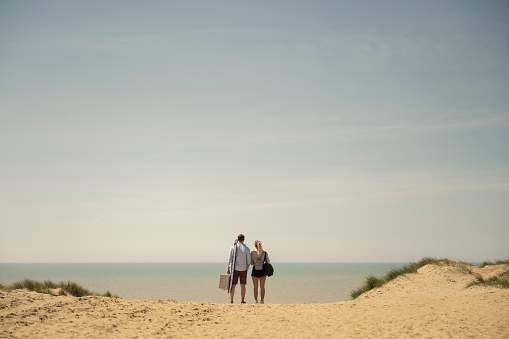 Mid distance rear view of casually dressed Caucasian couple in 20s and 30s carrying picnic basket and pausing at crest of sand dune to admire scenery.