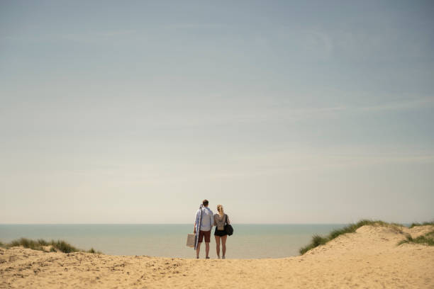 Man and woman on beach holiday enjoying ocean view Mid distance rear view of casually dressed Caucasian couple in 20s and 30s carrying picnic basket and pausing at crest of sand dune to admire scenery. mid distance stock pictures, royalty-free photos & images