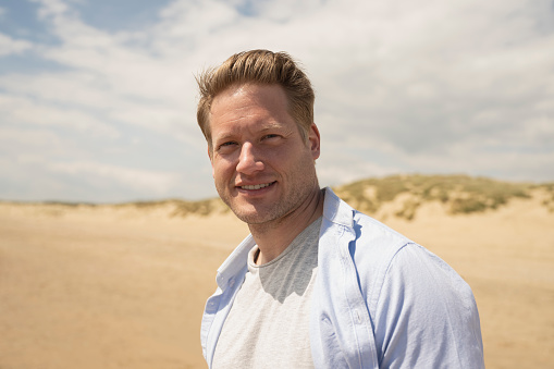 Head and shoulders view of blond man wearing casual clothing and pausing from walking near water’s edge to smile at camera with sand dunes in background.