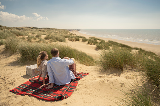 Rear perspective of Caucasian couple in 20s and 30s reclining on blanket with picnic basket at Camber Sands beach.