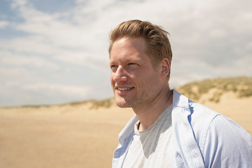 Close-up of mid 30s Caucasian man with short blond hair wearing casual clothing and looking away from camera while walking near water’s edge.