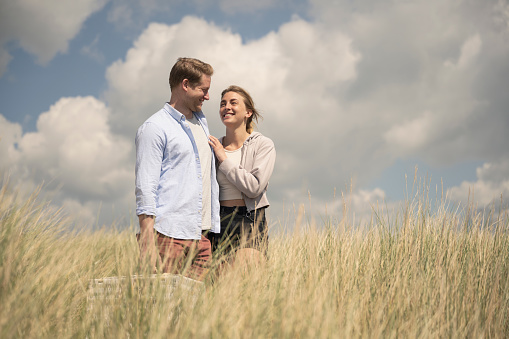 Caucasian man and woman in 20s and 30s smiling face to face as they walk across sand dune with picnic basket for day of leisure.