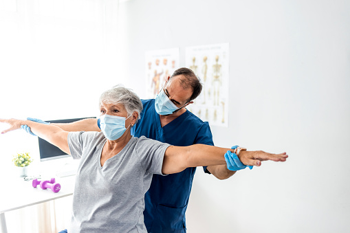 Doctor physiotherapist consulting with patient about shoulder muscle pain problems,  wearing protective mask. Physical therapy diagnosing concept.