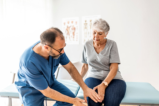 Photo of senior woman having some knee pain. She's at doctor's office having medical examination by a male doctor. The doctor is touching the sensitive area and trying to determine the cause of pain.