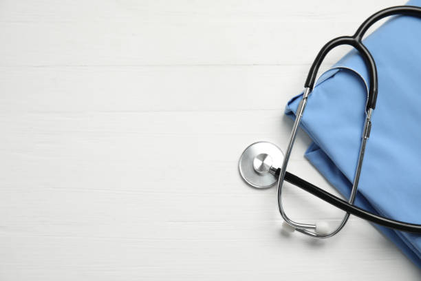 stetoscopio e uniforme medica su sfondo bianco in legno, posa piatta. spazio per il testo - camice da medico foto e immagini stock