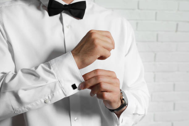 Stylish man putting on cufflink near white brick wall, closeup