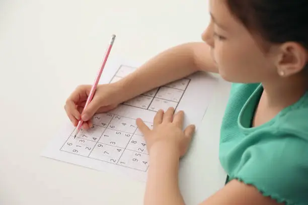 Little girl solving sudoku puzzle at white table, closeup