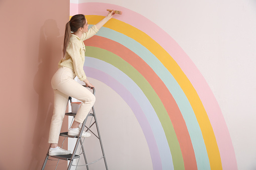 Young woman painting rainbow on white wall indoors, space for text