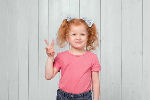 Portrait of cute carefree ginger little girl 4-6 years old, showing peace v-sign gesture. Studio shot, light wooden background