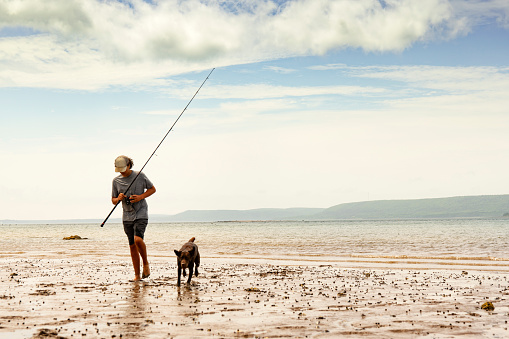 A young fisherman, surf fishing with his Chocolate Labrador Puppy on a beautiful, rugged, Annapolis Basin beach in Nova Scotia.
