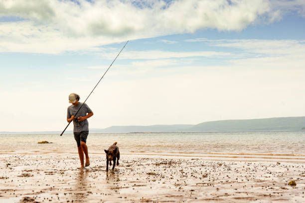 A teenaged surf-fisherman on a beautiful beach fishing with his dog A young fisherman, surf fishing with his Chocolate Labrador Puppy on a beautiful, rugged, Annapolis Basin beach in Nova Scotia. sea fishing stock pictures, royalty-free photos & images