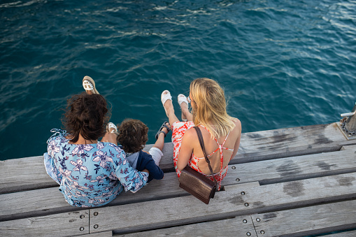 Little boy enjoying summer day on wooden dock with his mother and aunt, during coronavirus pandemic after reopening borders