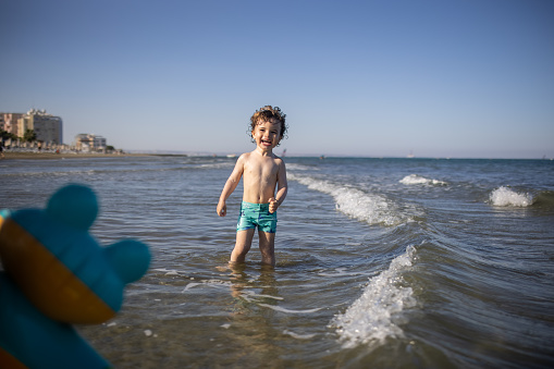 Happy little boy having fun in sea during beach holiday, during coronavirus pandemic after reopening borders