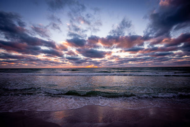 The image captures a scenic coastal sunset, showing an orange sky blending into the calm ocean. The waves roll gently onto the sandy shore, mirroring the vibrant colors of the sky.