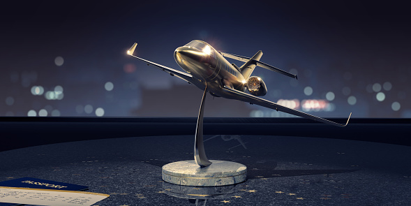 A gold an silver model of a generic private jet with a marble base on a dark granite table, surrounded by gold foil star shapes close to a passport and first class boarding pass. The model is near a window with bokeh from lights of nearby buildings.