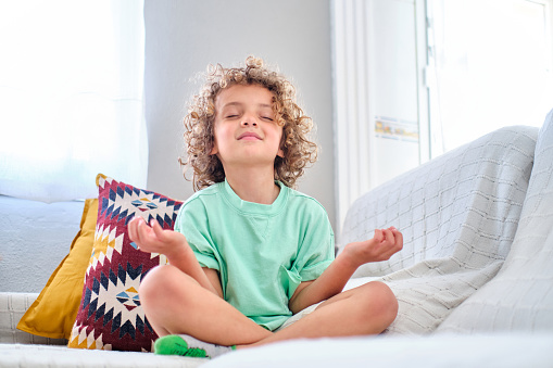 child practicing meditation sitting on the sofa at home in lotus pose