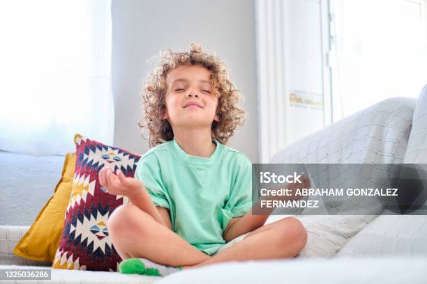 Child Practicing Meditation Sitting On The Sofa At Home In Lotus Pose Stock Photo - Download Image Now