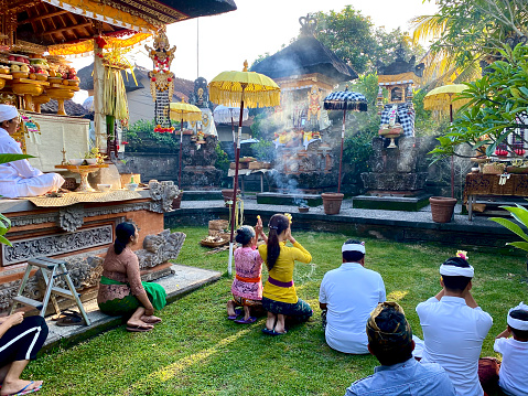 Horizontal photo of a group of people sitting on the grass in the decorated family temple area, in ceremony, wearing traditional clothing. The priest can be seen sitting on a raised platform, leading the ceremony.