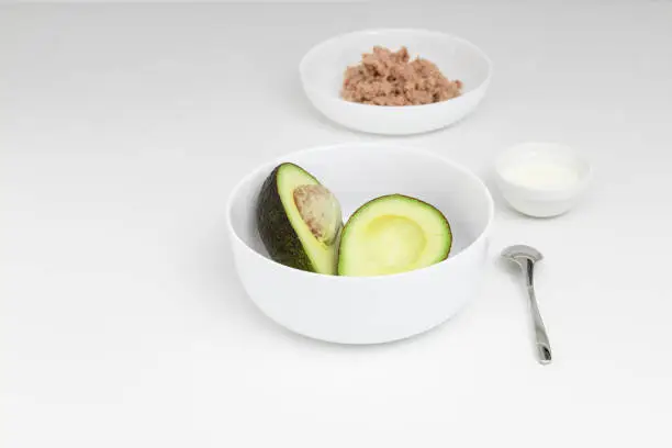 Sliced avocado on the white plate in the white-color kitchen. Yogurt and tuna on the plate are on the background. Step-by-step recipe.