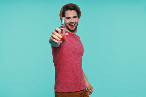 Handsome young man in casual clothing holding bottle with drink and smiling while standing against blue background