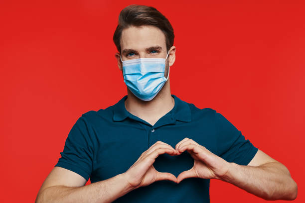 istock Handsome young man in protective face mask looking at camera and gesturing heart shape 1325051233