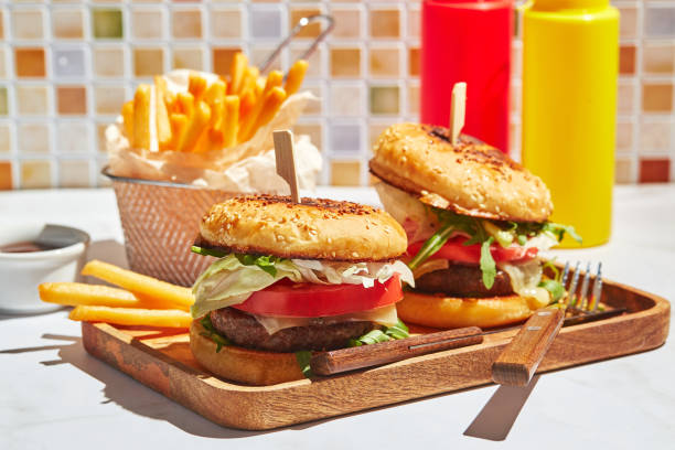 delicious homemade burgers on wooden tray in bright sunlight. burgers with veal cutlet, pamidor, cheese, red onion, lettuce, arugula and fries. fast food concept - sesame bun american culture cheddar imagens e fotografias de stock