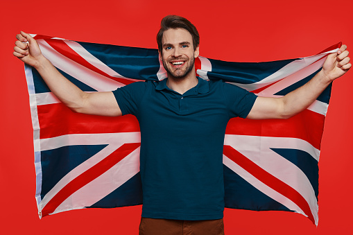Handsome young man in casual wear carrying British flag and smiling while standing against red background