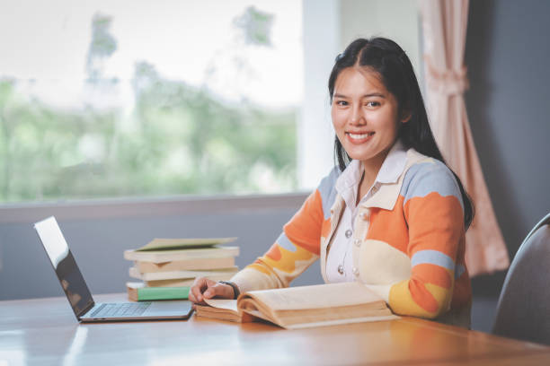 Young Asian woman college student in student uniform studying, reading a book, laptop in university or college library. Youth girl student and tutoring education with a technological learning concept stock photo