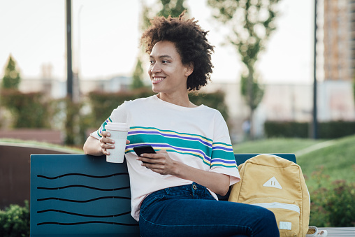 Portrait of a happy, young African American woman enjoying coffee in the park and using a smartphone.