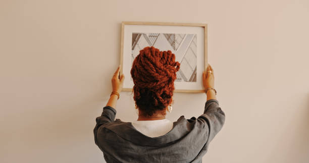 shot of a unrecognizable female hanging a painting at home - hangen stockfoto's en -beelden