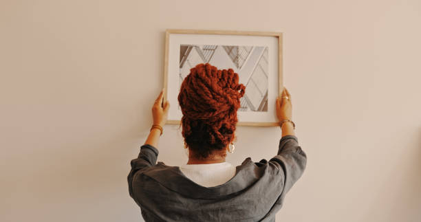 shot of a unrecognizable female hanging a painting at home - home decorating fotos imagens e fotografias de stock