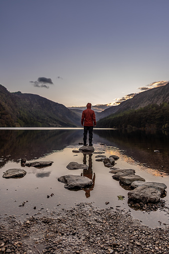 Man in orange jacket standing on stone in the middle of Glendalough Lake in Wicklow Ireland