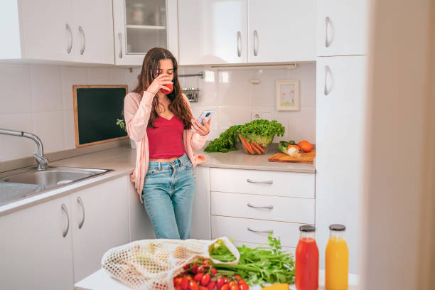 건강한 젊은 여자 마시는 토마토 주스와 그녀의 부엌에서 그녀의 휴대 전화를 사용 하 여 - vegetable juice juice carrot tomato 뉴스 사진 이미지